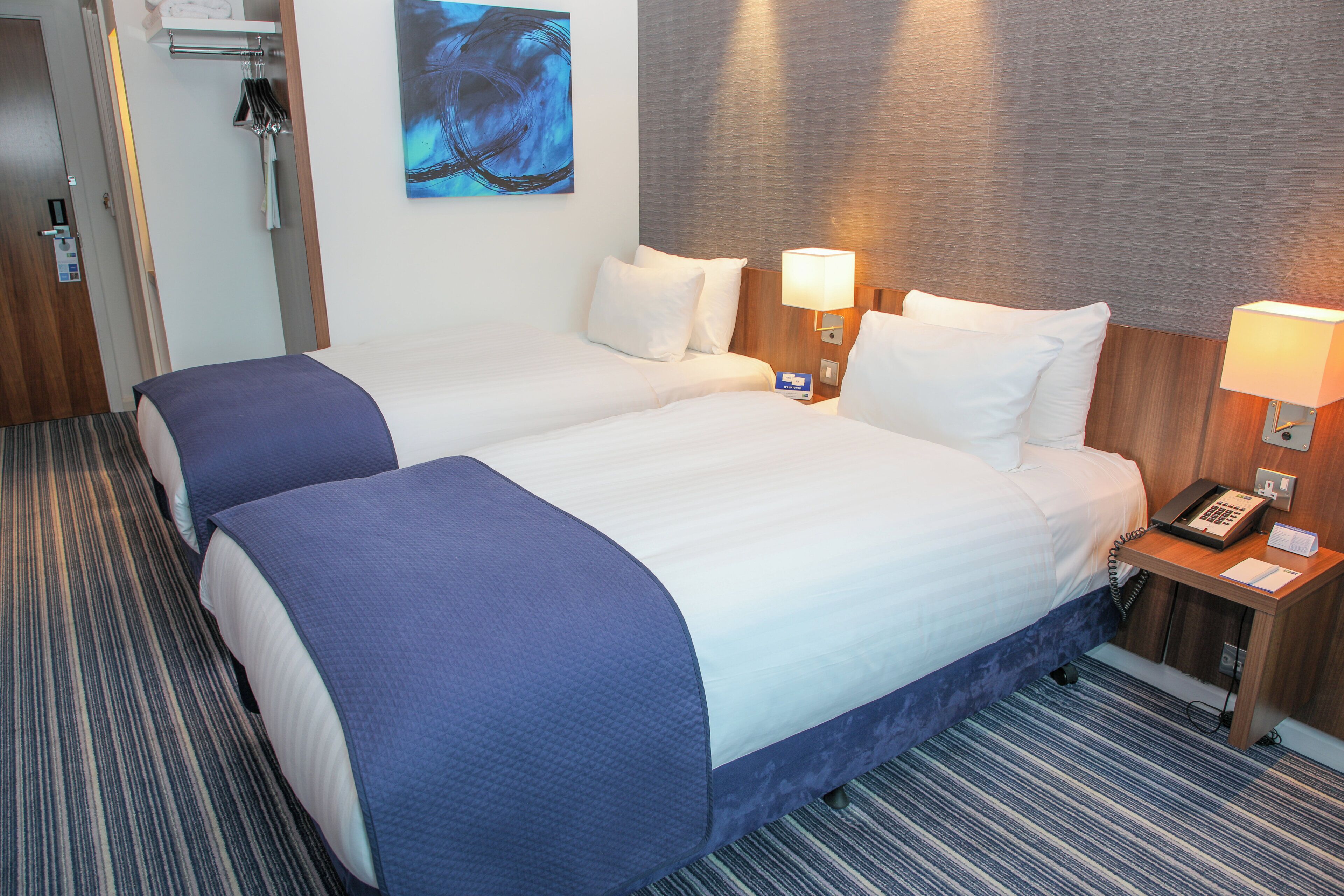 A hotel room with two beds, a wooden headboard, a bedside table with a lamp, and a painting on the wall.