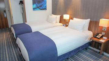 A hotel room with two beds, a wooden headboard, a bedside table with a lamp, and a painting on the wall.