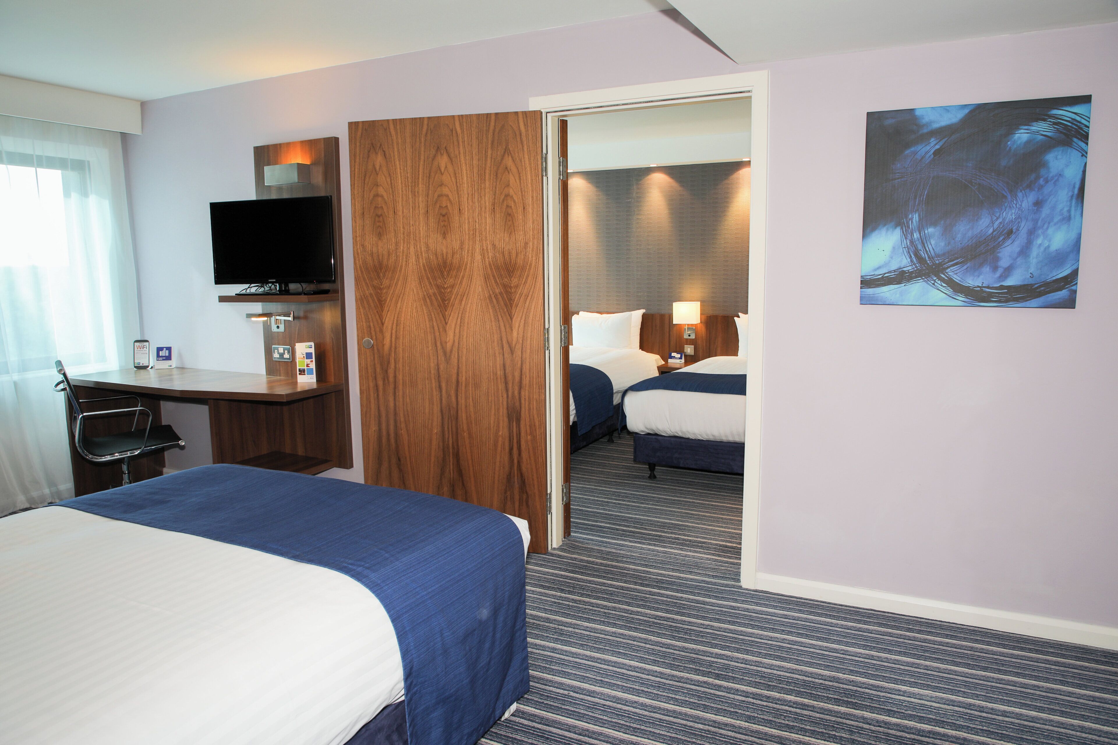 Une chambre d’hôtel comprenant un lit, un bureau avec un téléviseur à écran plat, une armoire en bois et une porte donnant sur une autre pièce.