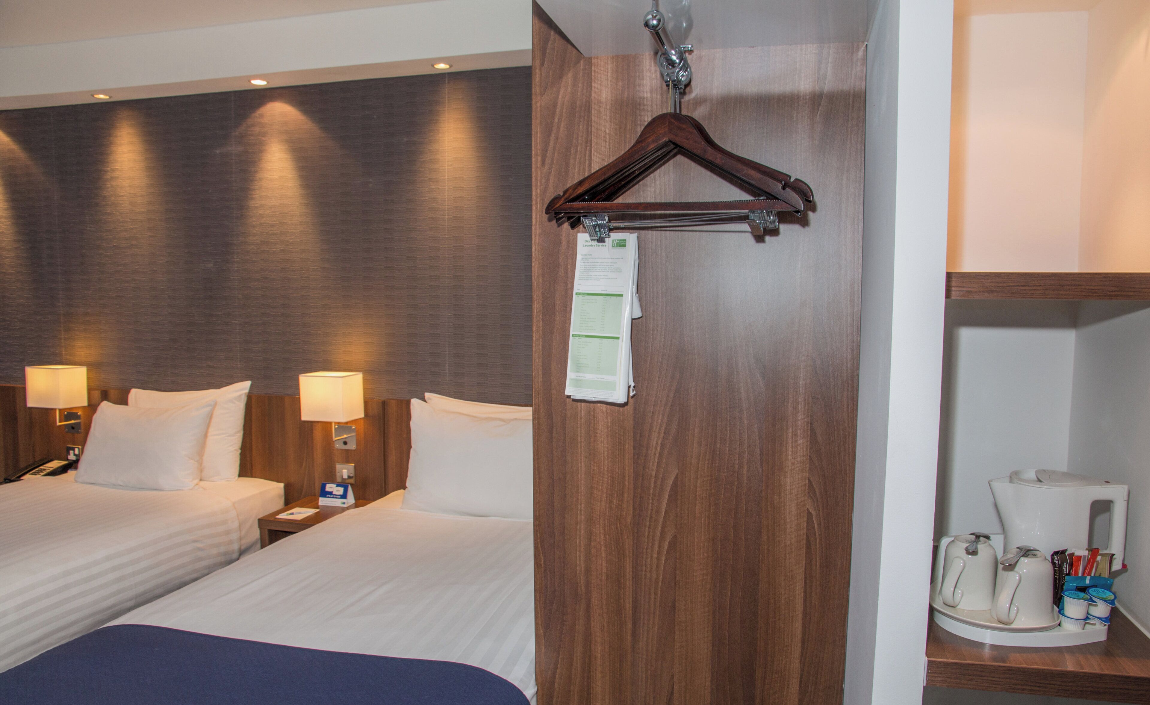 A hotel room with two beds, a wooden headboard, a hanging coat rack, a bedside table with a lamp, and a coffee maker on a tray.