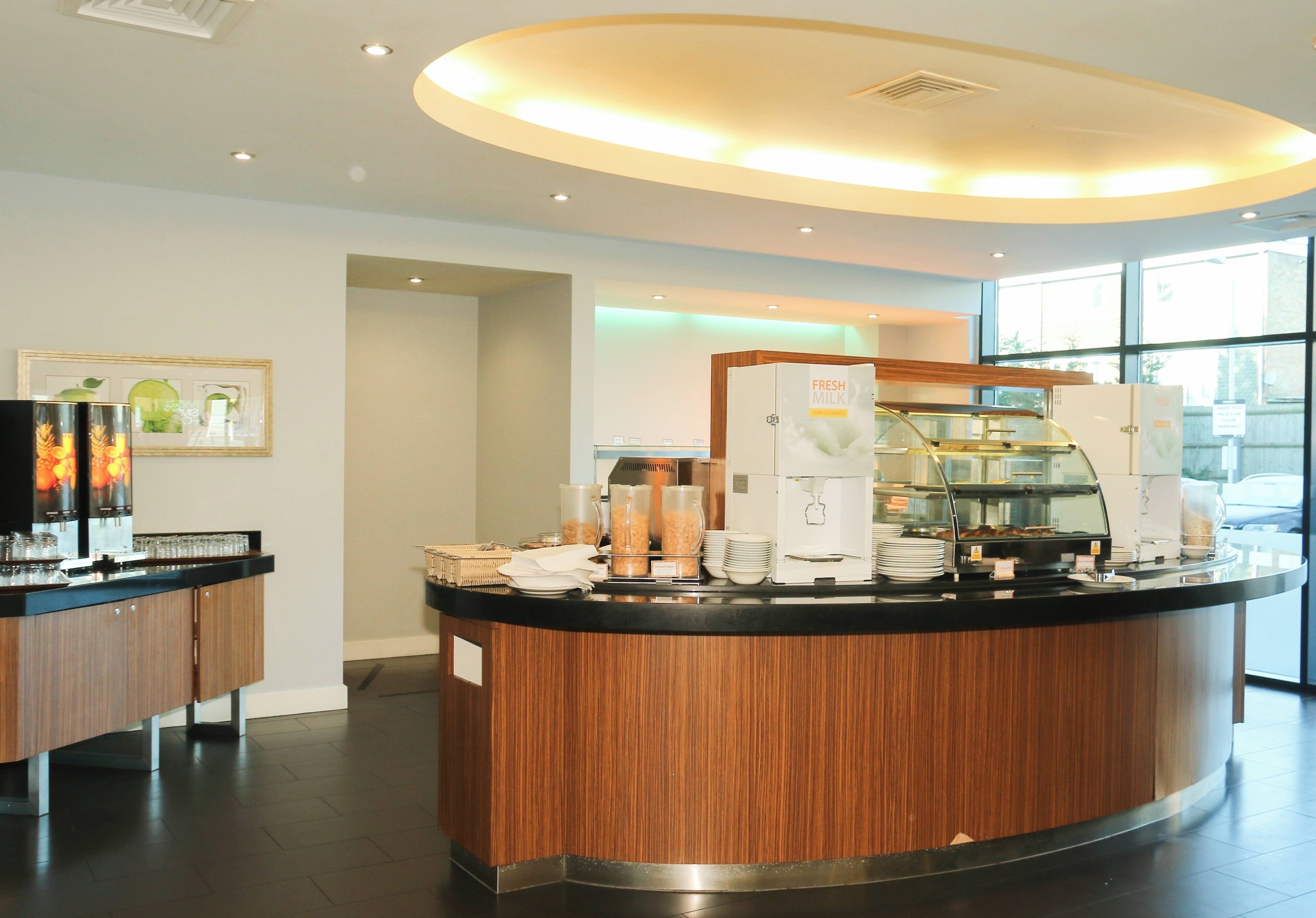 Una barra de desayuno moderna con detalles de madera, equipada con una máquina de café, dispensador de leche fresca y diversas opciones para el desayuno.