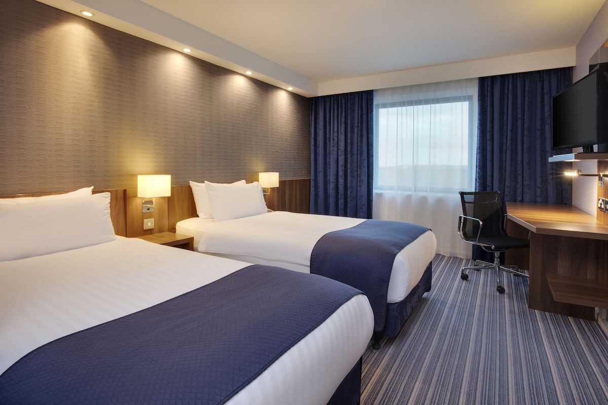 A hotel room with two beds, a desk, a chair, a television, and a large window with curtains.