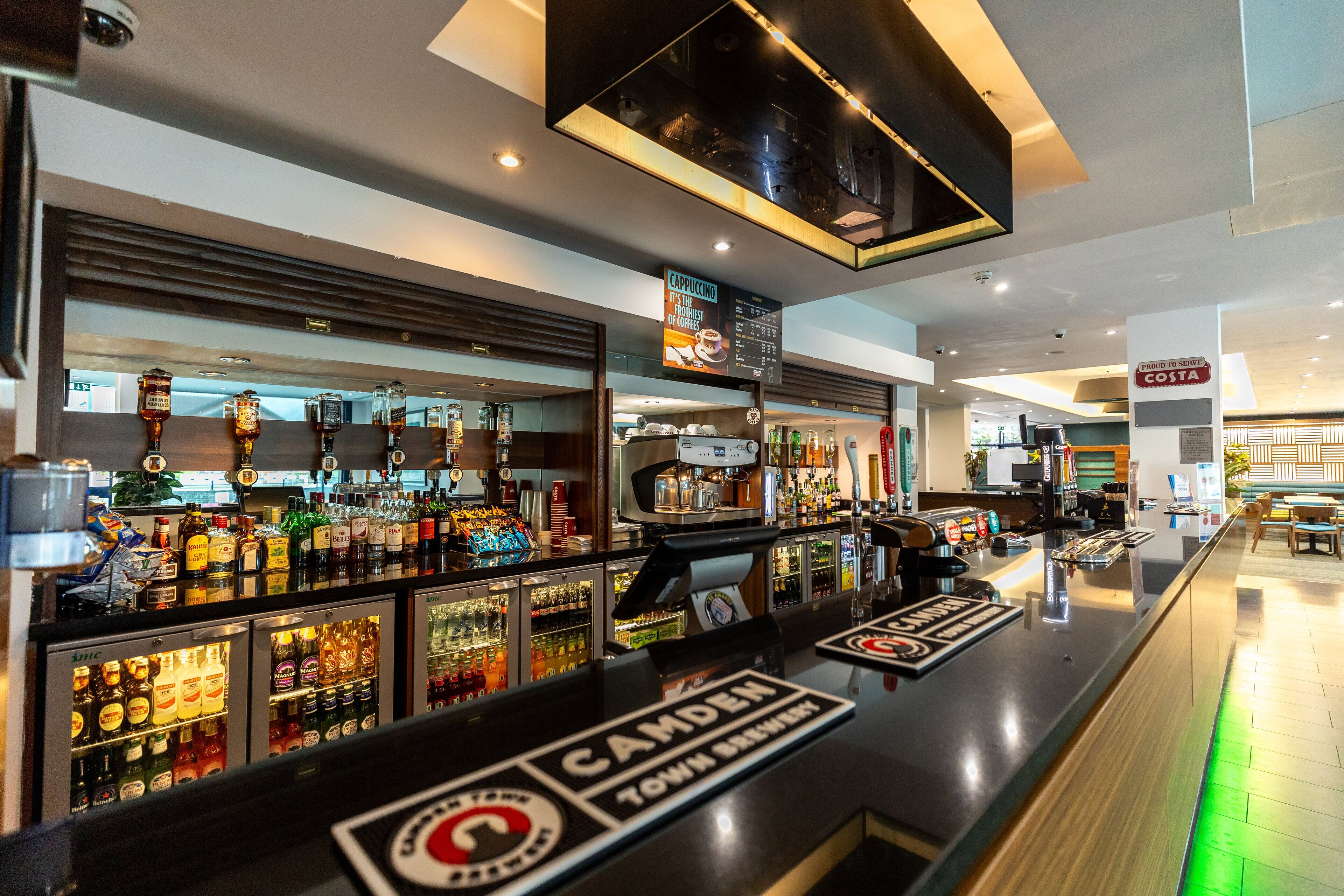 A bar area with a modern design, including a counter with a sign reading "CAMDEN TOWN BREWERY" and a menu board above it.