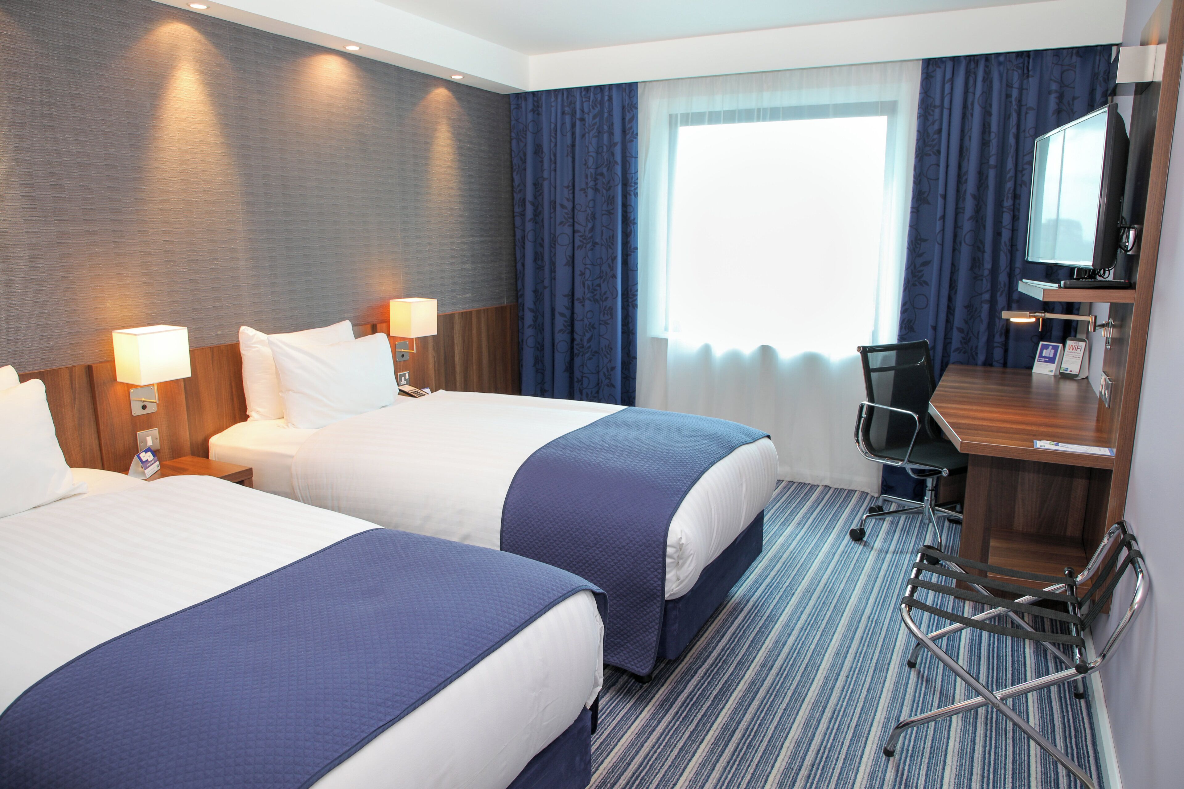 Een hotelkamer met twee bedden, een bureau, een stoel en een groot raam met gordijnen.