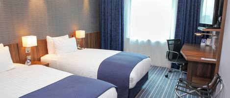 Ein Hotelzimmer mit zwei Betten, einem Schreibtisch, einem Stuhl und einem großen Fenster mit Vorhängen.