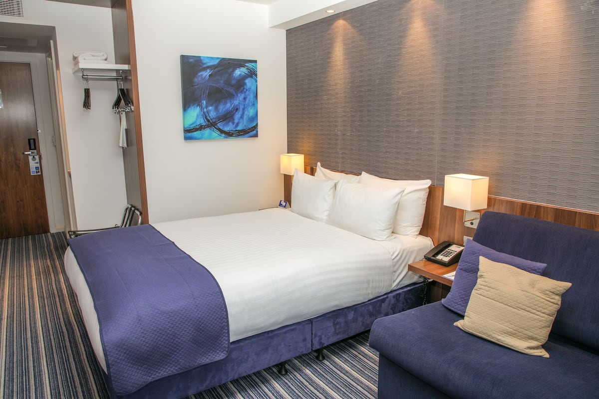 A hotel room with a bed, a sofa, a painting, a lamp, and a telephone.