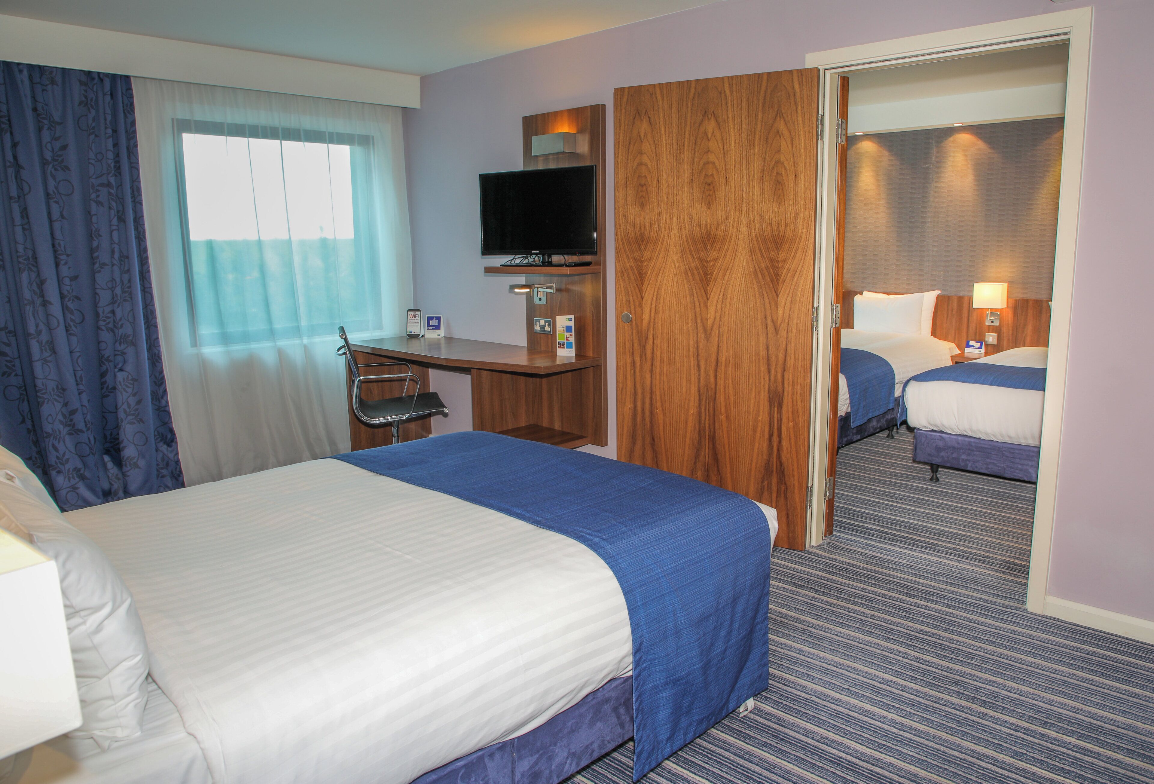 Quarto de hotel com cama, escrivaninha, televisão e espelho.