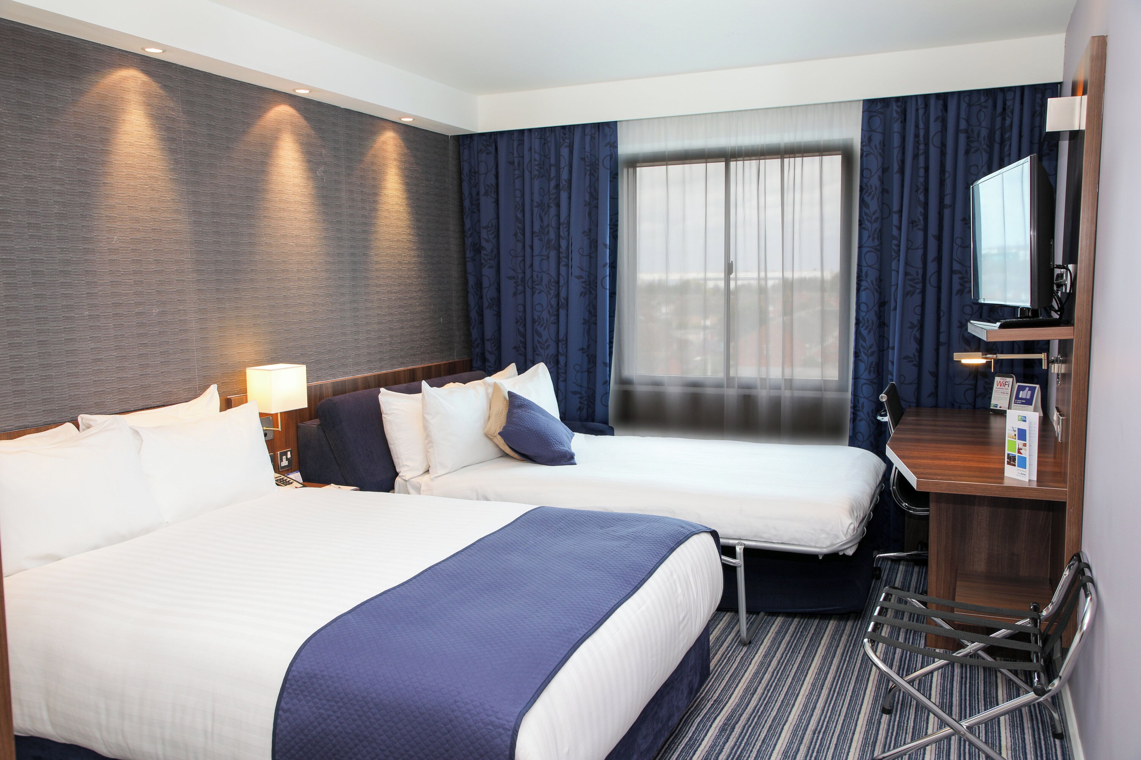 A hotel room with two beds, a desk, a chair, a TV, and a window with curtains.