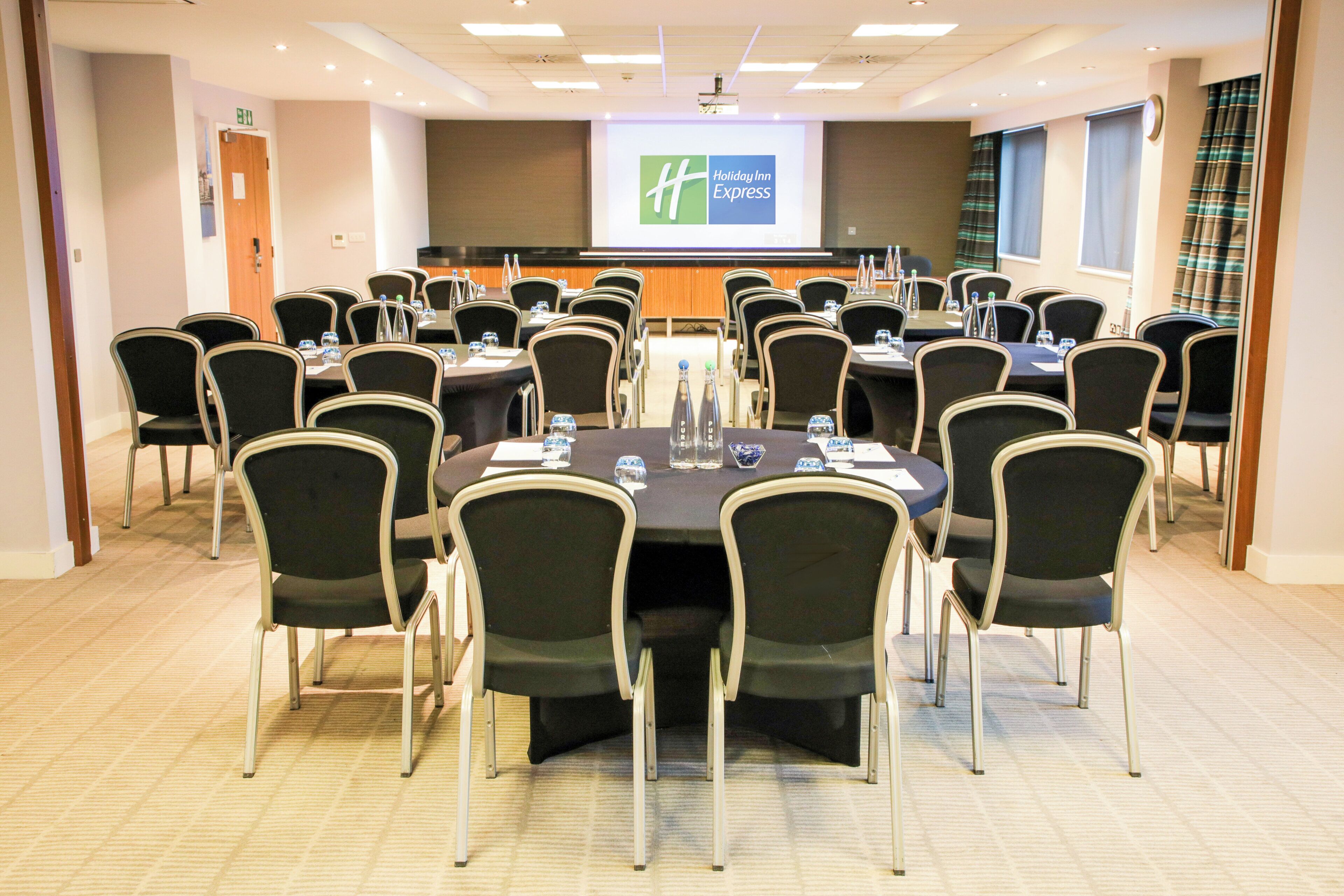 A conference room with multiple tables and chairs, equipped with water bottles and nameplates, and a presentation screen in the background.