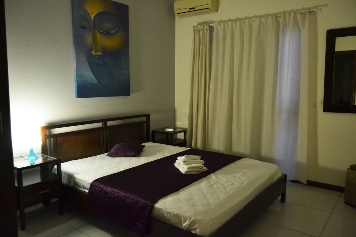 A neatly made bed with a headboard, a bedside table with a lamp, and a painting on the wall.