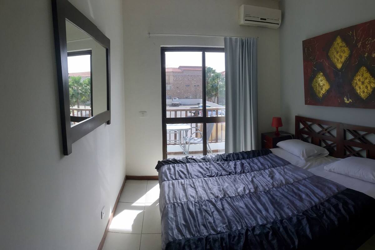 A bedroom with a bed, a nightstand, a red lamp, a wall-mounted air conditioner, and a view of the outside balcony.