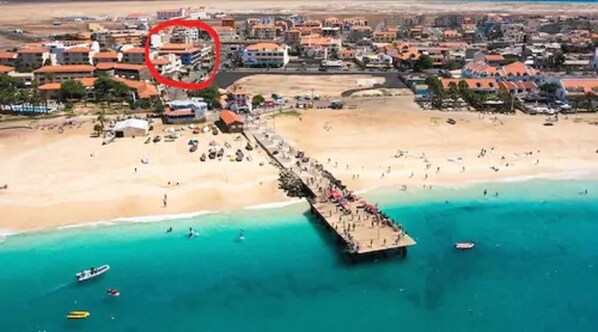 An aerial view of a coastal area with a pier extending into turquoise waters, surrounded by a beach populated with people and boats.