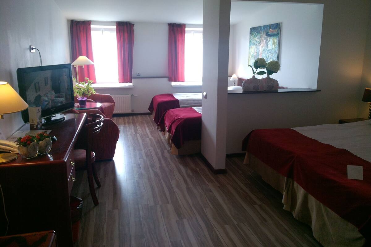 Habitación de hotel con piso de madera, un escritorio con lámpara, un televisor, dos camas con ropa de cama roja, una ventana con cortinas rojas y un cuadro en la pared.