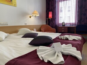 A hotel room with a bed, pillows, a desk, a chair, a lamp, and a window with curtains.