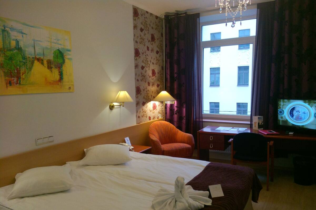 Una habitación de hotel con cama, escritorio con computadora, una silla y ventana con cortinas.