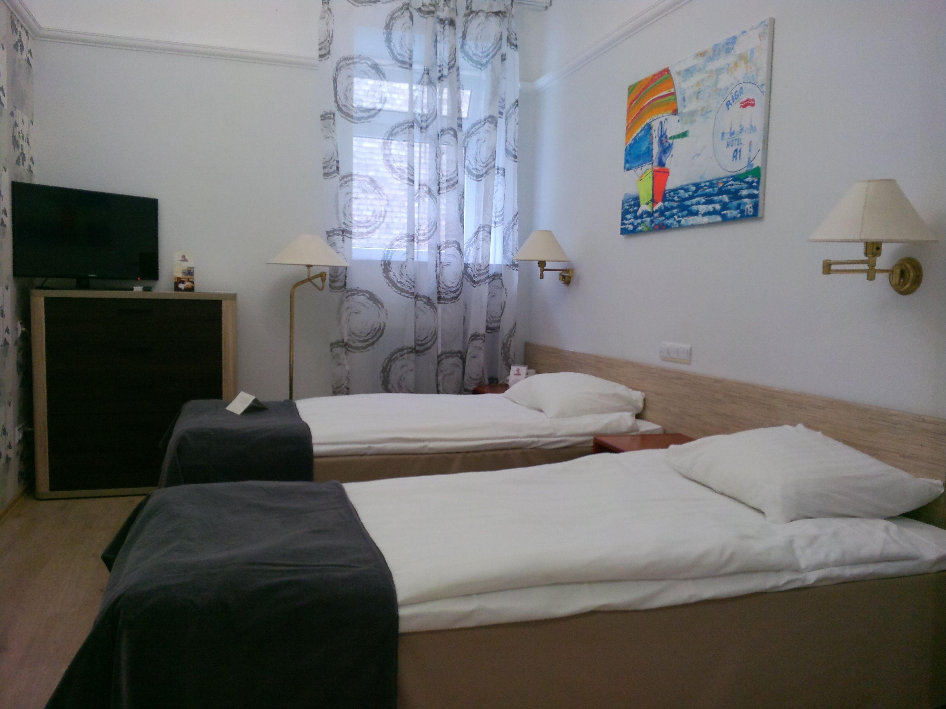 Quarto de hotel com duas camas de solteiro, uma televisão em um suporte, duas luminárias e um quadro na parede.