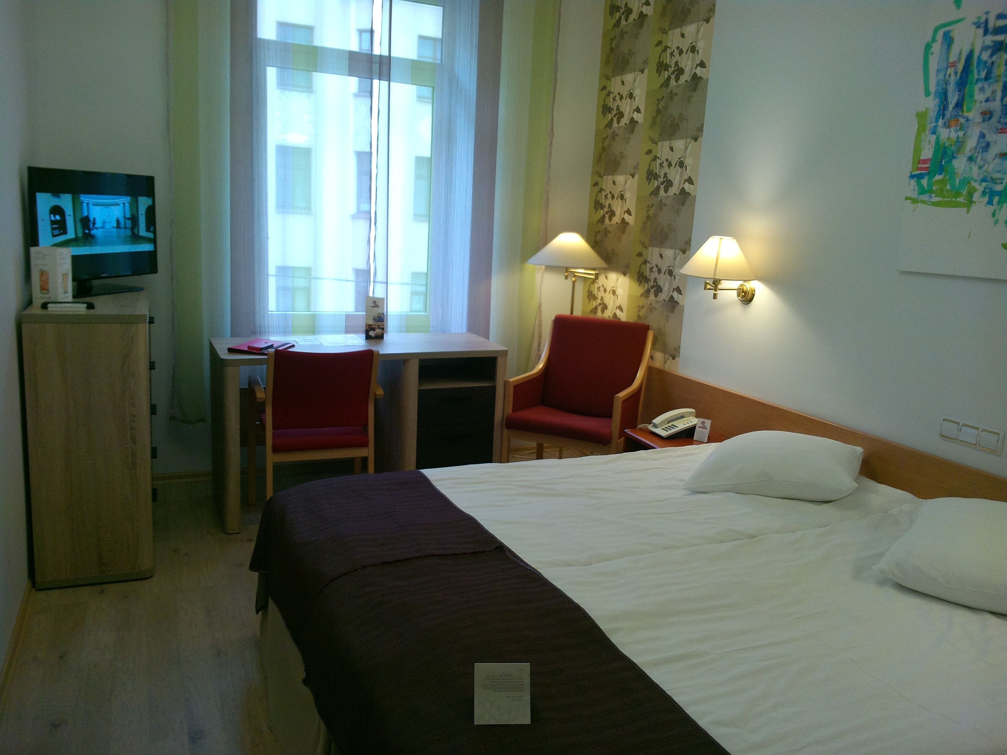 Hotellihuone, jossa on sänky, työpöytä, kaksi tuolia, televisio ja lamput.