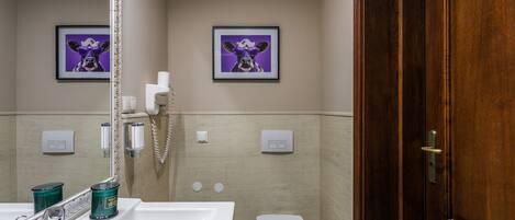 Une salle de bain avec un lavabo blanc, un miroir et des toilettes. Deux tableaux encadrés ornent le mur, ainsi qu’une porte en bois et un porte-serviettes.