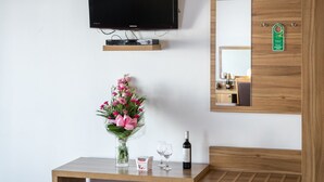 A modern room with a wooden ceiling, a flat-screen TV mounted on the wall, a wooden bench, a small table with a vase of flowers, and a mirror with a green sign. - Aparthotel Pergamin (Kraków)