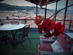 A balcony with a table and chairs, a patterned sofa, and a view of a cityscape with buildings and hills. - San Francisco Plaza Hotel (Cusco)