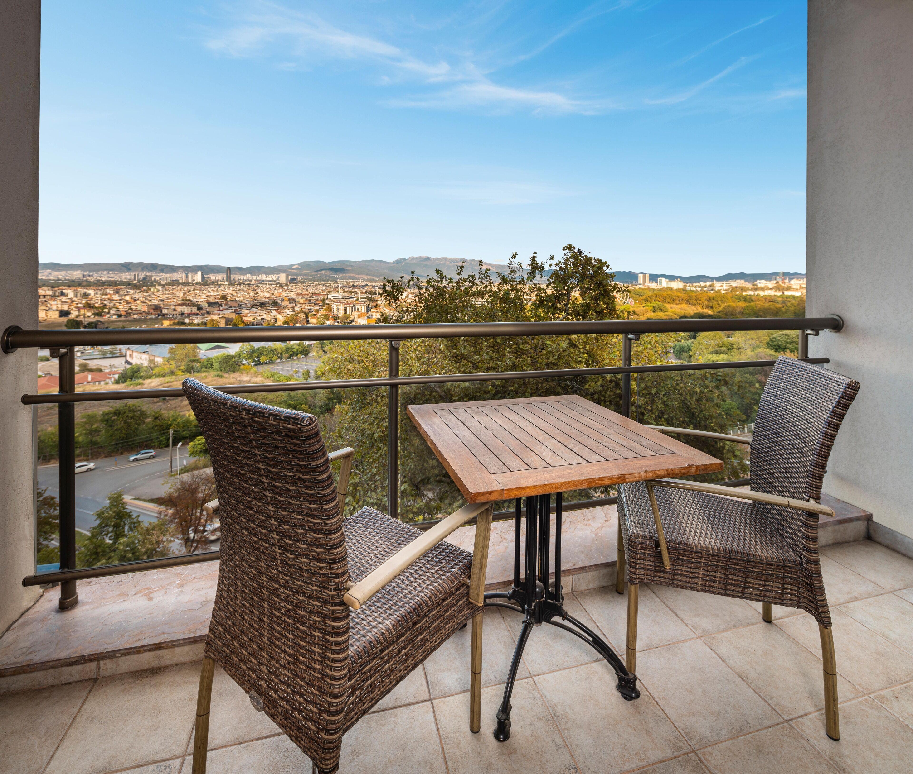 A balcony with wicker chairs and a wooden table, offering a view of a cityscape and mountains.