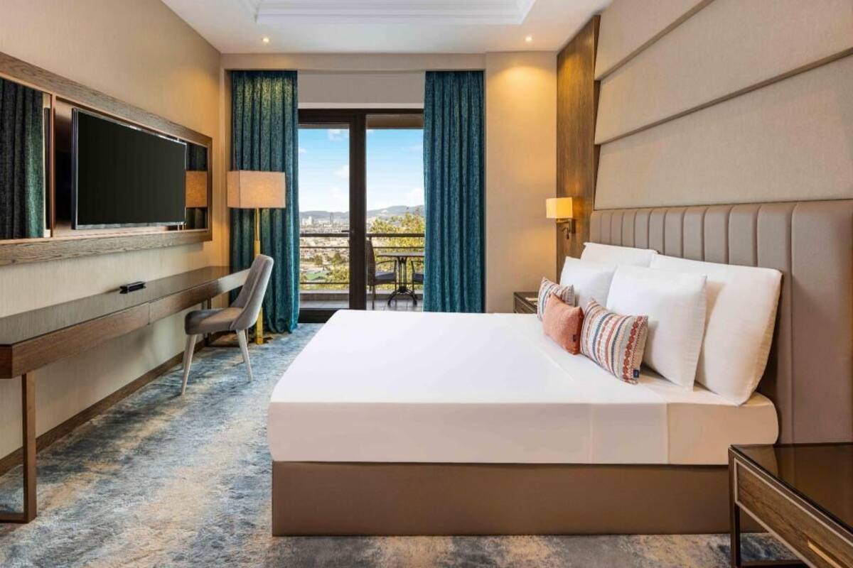 A hotel room with a large bed, a desk, a TV, and a view of the city.