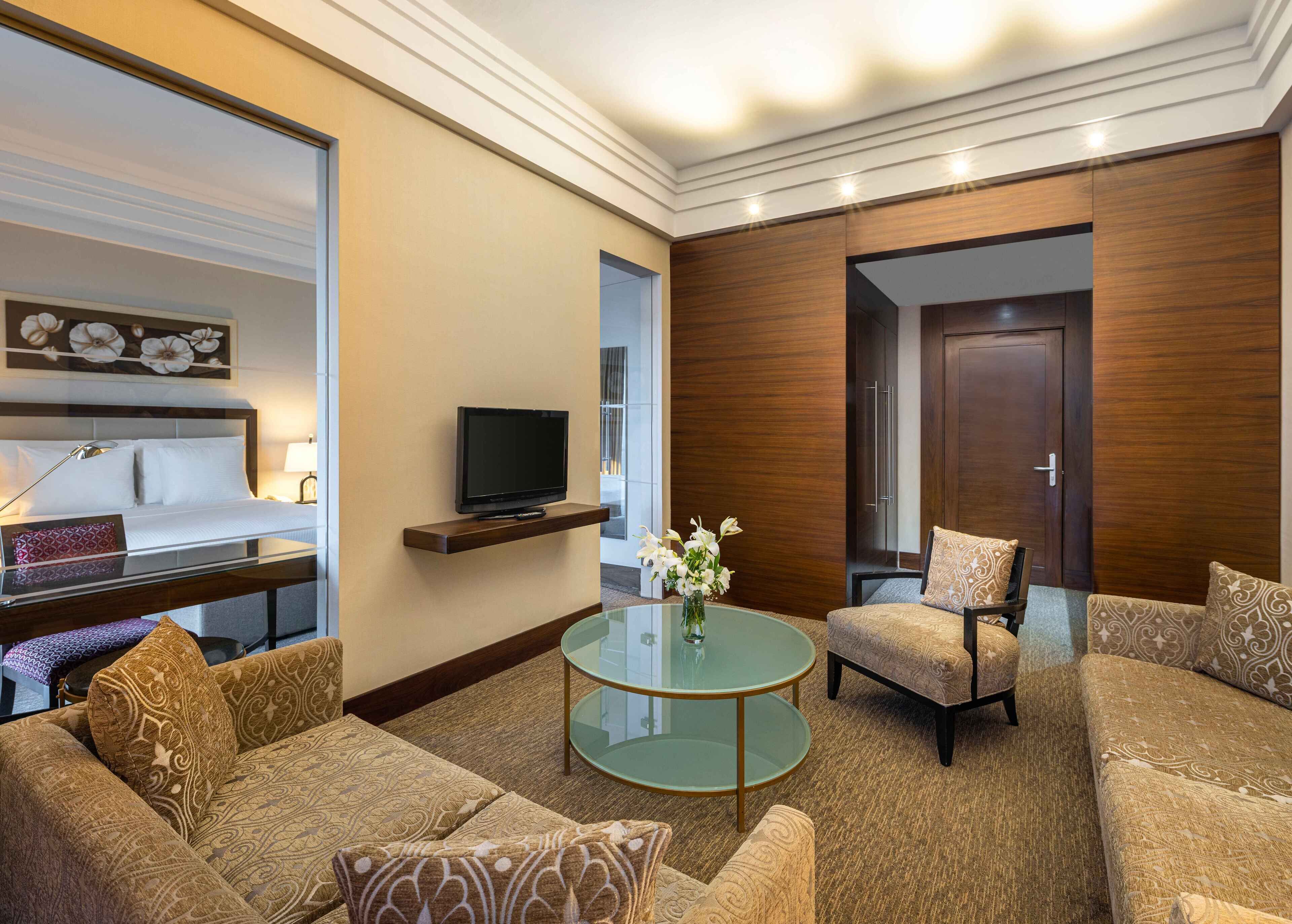 A hotel room with a bed, a TV on a shelf, a glass coffee table, a sofa, and a chair.