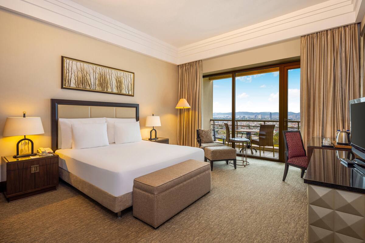 A hotel room with a large bed, a desk with a TV, a chair, a small table, and a view of the city through large windows.