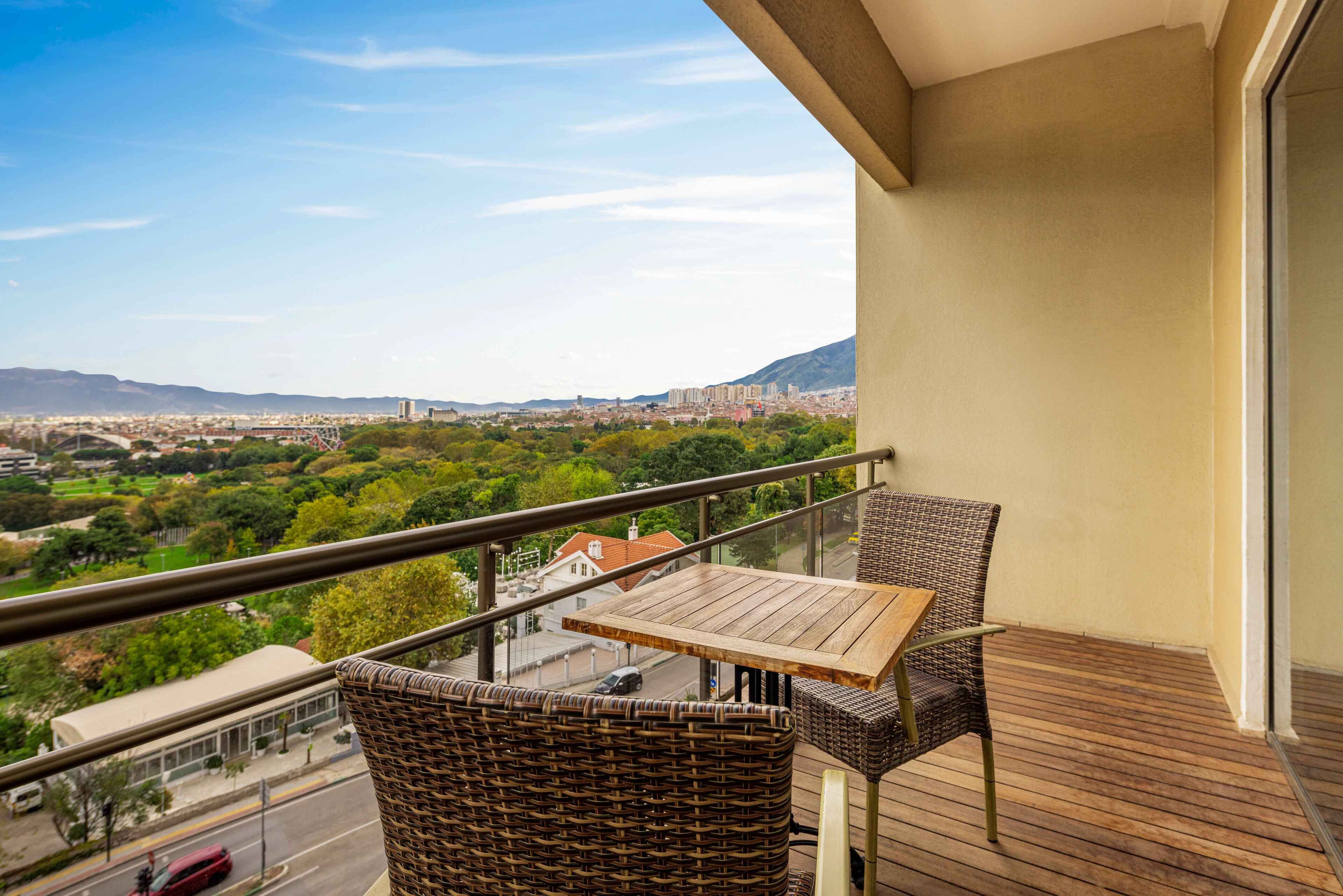 A balcony with wicker furniture and a wooden table, overlooking a cityscape and greenery.