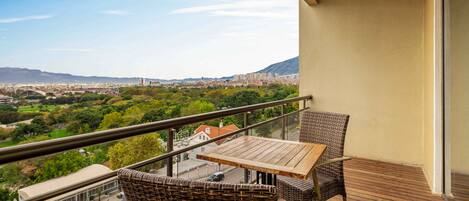 A balcony with wicker furniture and a wooden table, overlooking a cityscape and greenery.