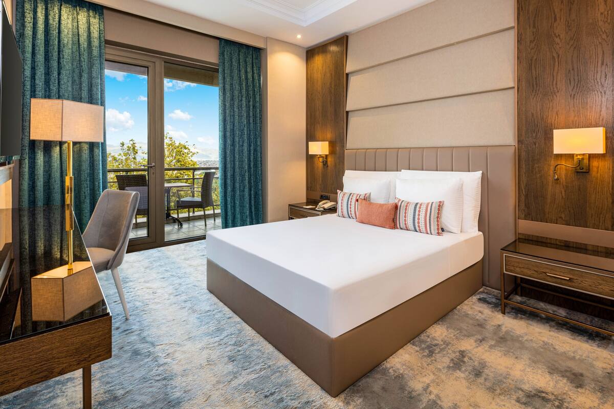 A modern hotel room with a large bed, a desk with a lamp, a chair, and a view of the outdoors.