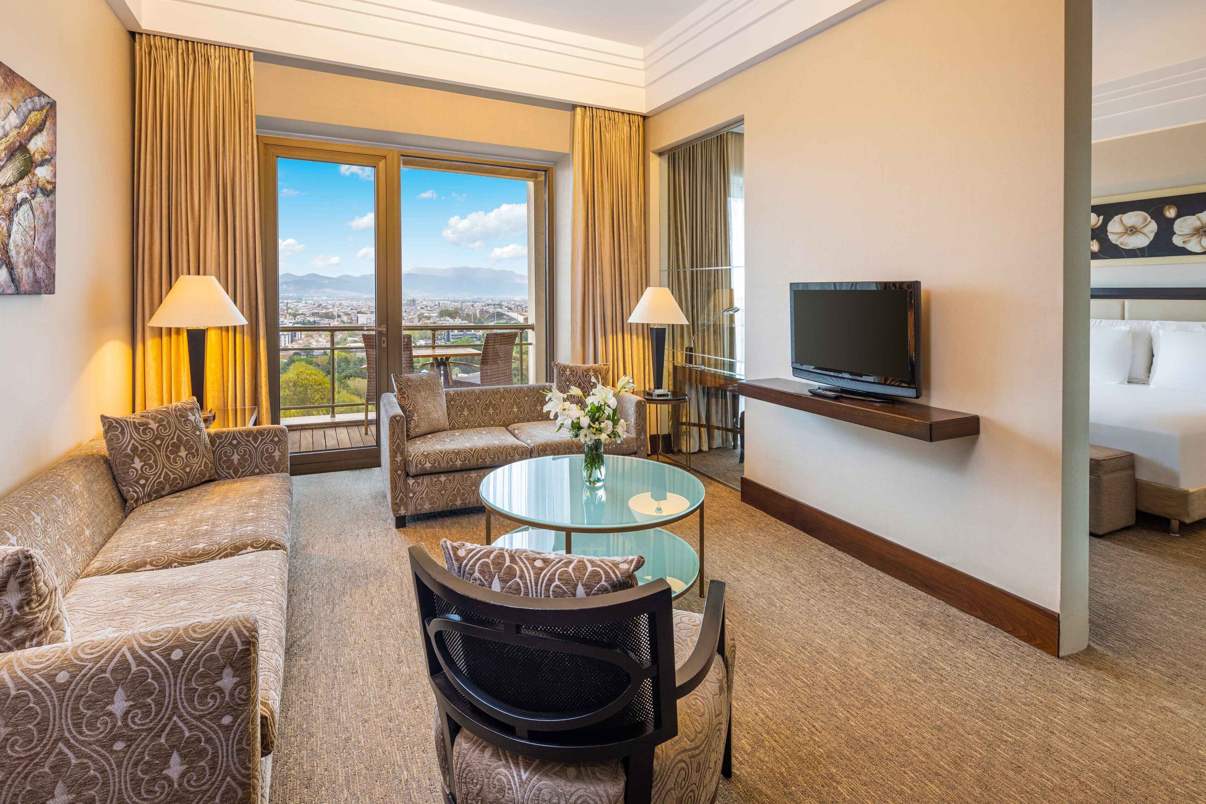 A hotel room with a sofa, armchair, and a glass table with a vase of flowers. There is a TV mounted on the wall and a balcony with a city view.