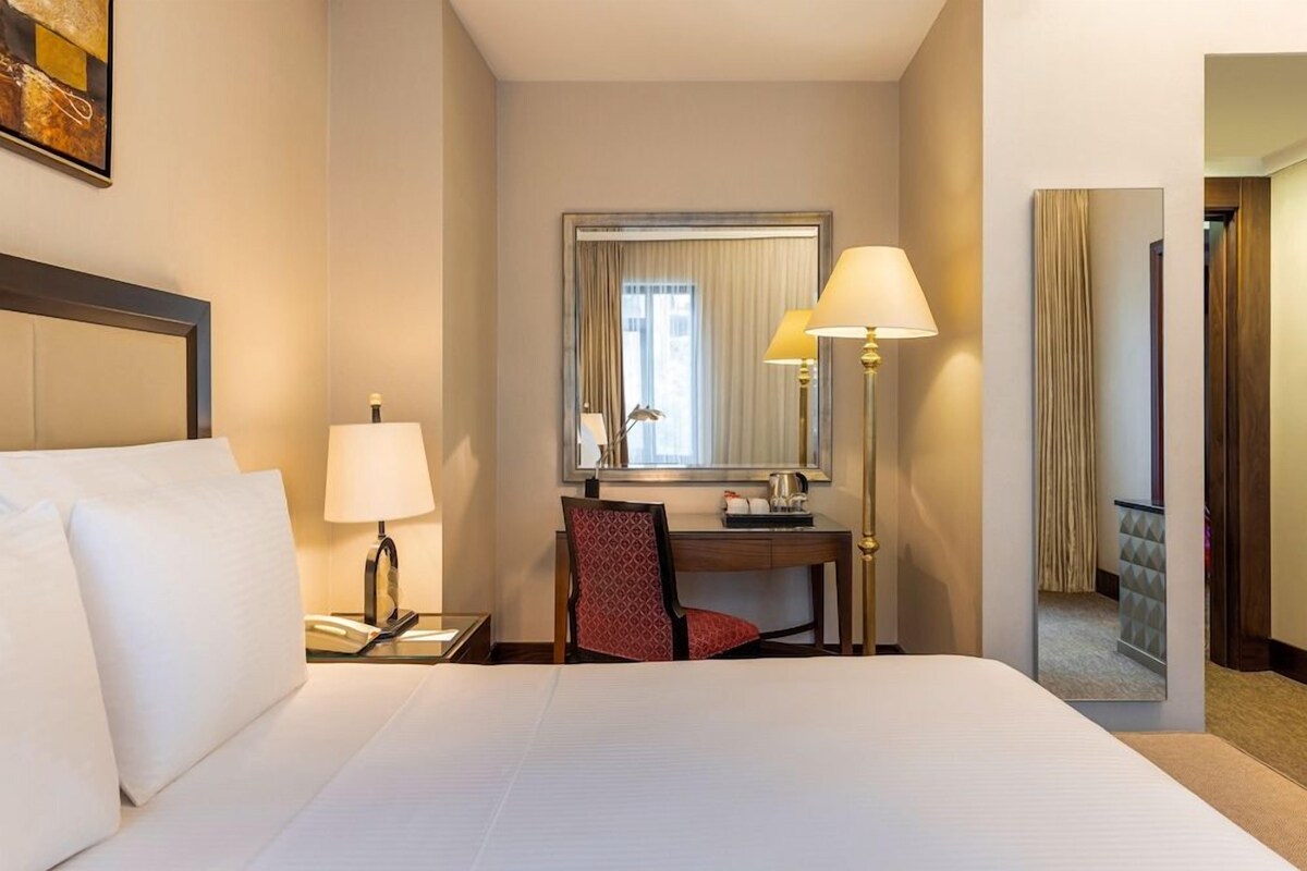 A hotel room with a bed, a desk, a chair, a lamp, and a mirror.