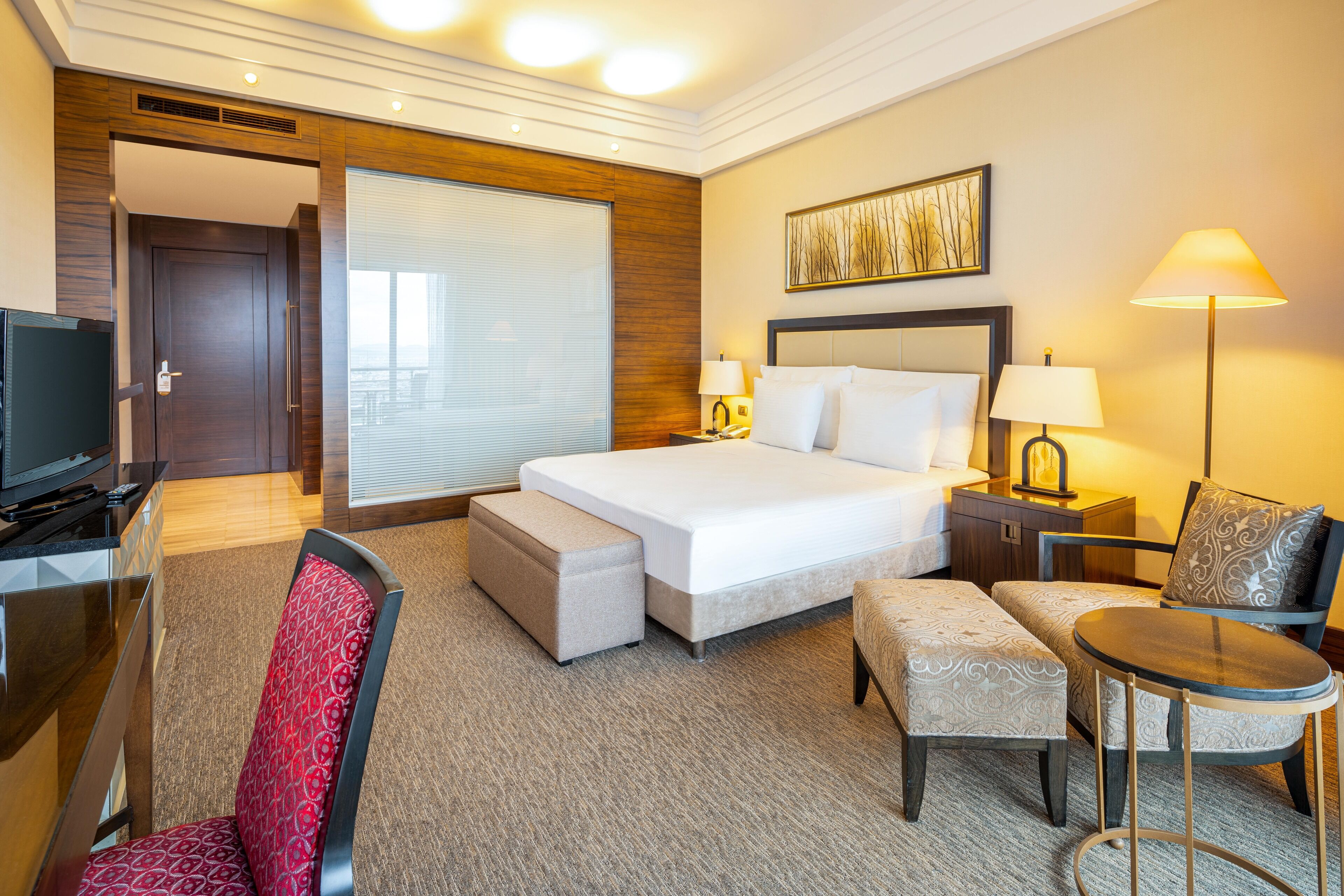 A hotel room with a large bed, a desk with a TV, a chair, a small table, and a lamp.