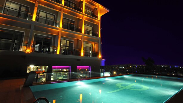 Un hotel moderno con piscina infinita en la azotea, varios balcones y vistas a la ciudad de noche.