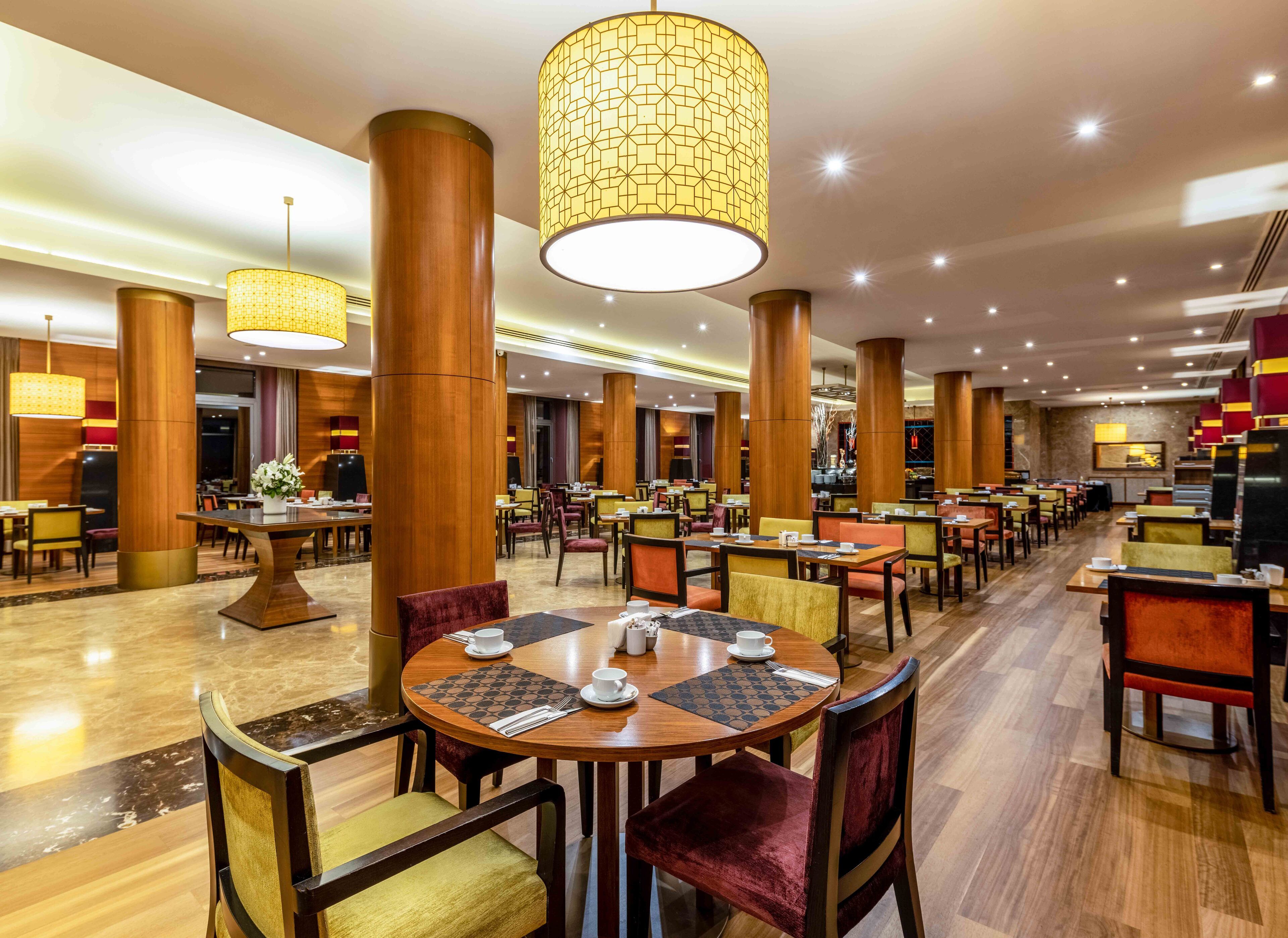 Un comedor de hotel espacioso con varias mesas y sillas redondas, altos pilares de madera y lámparas colgantes.