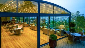 Un área de descanso al aire libre, moderna, con piso de madera, mobiliario de mimbre y amplios ventanales que ofrecen vistas de la ciudad.