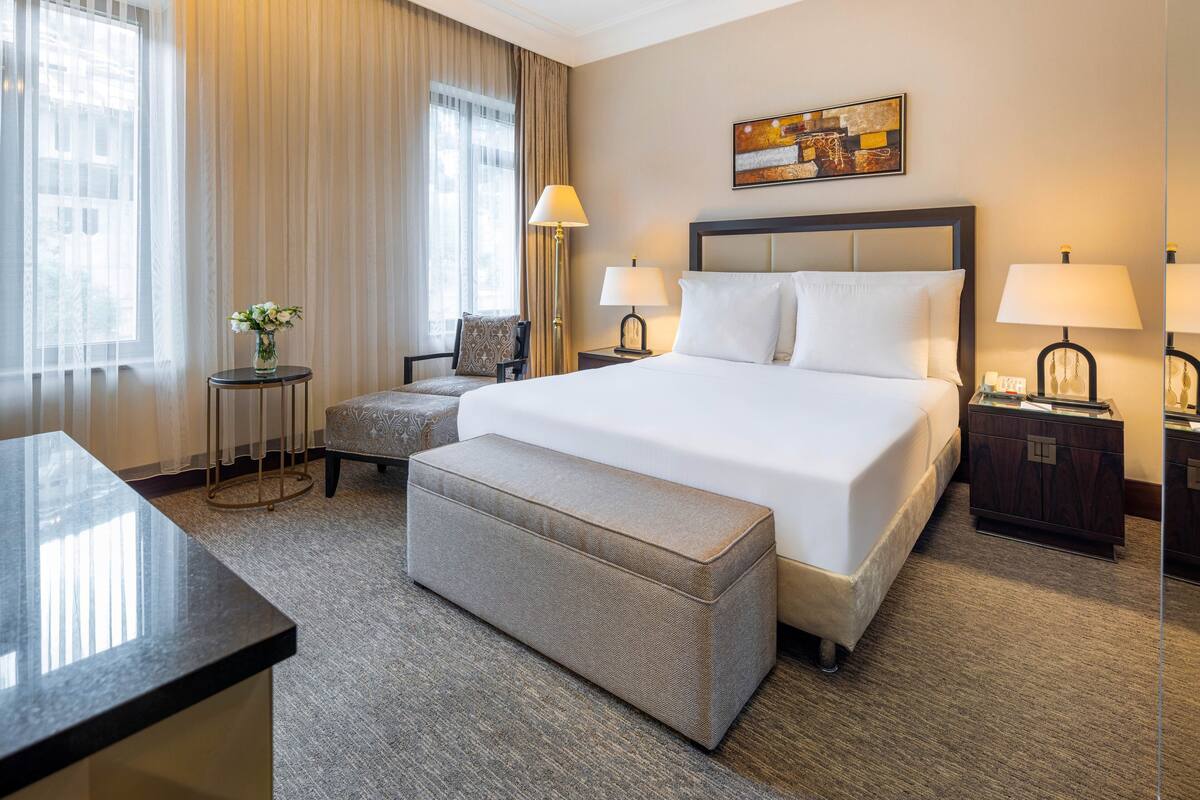A hotel room with a large bed, two bedside tables, a chair, a small table with a vase, and a large window with curtains.