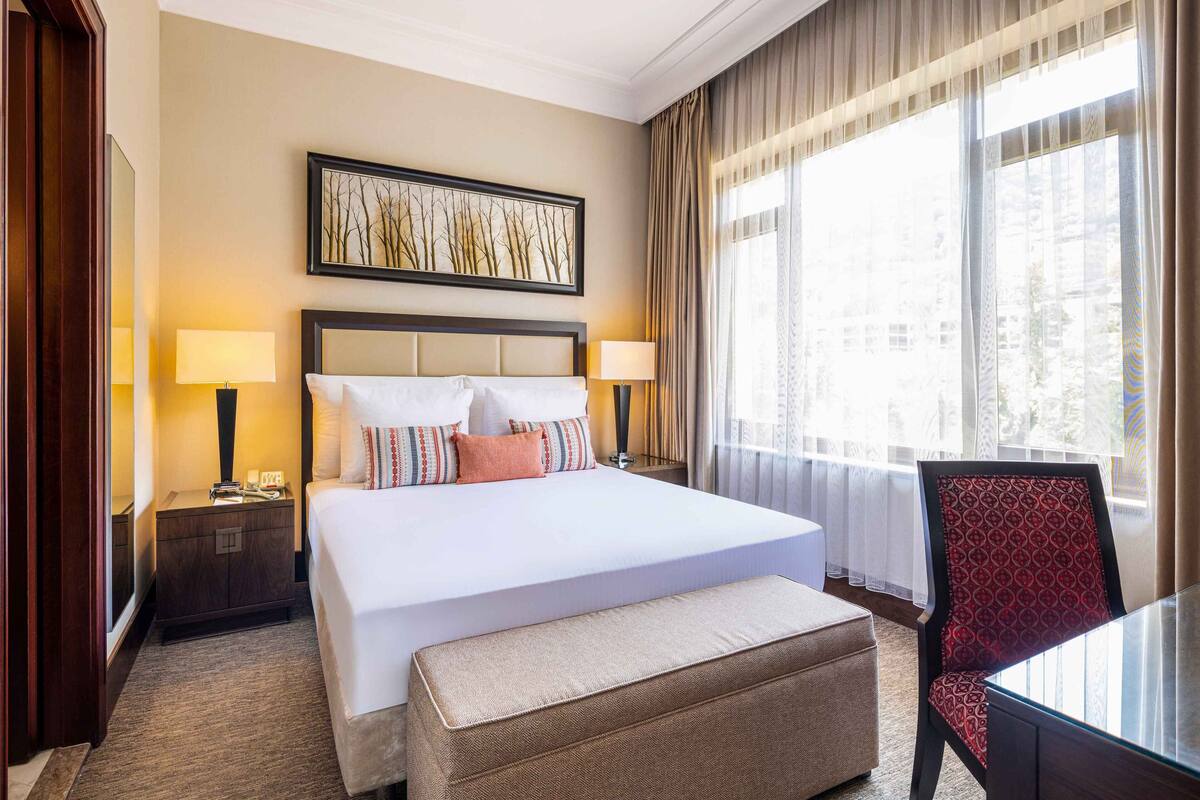 A hotel room with a large bed, two bedside tables with lamps, a large window with curtains, and a view of buildings outside.