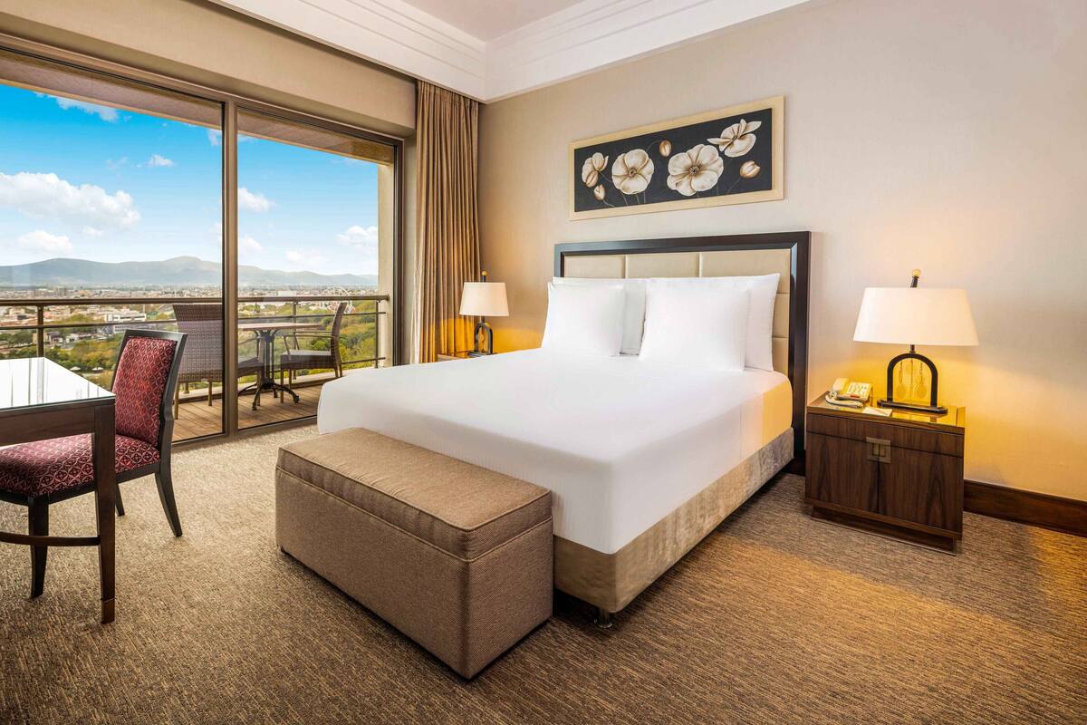 A hotel room with a large bed, two bedside tables, a desk with chairs, a balcony with a view of the city, and a painting on the wall.