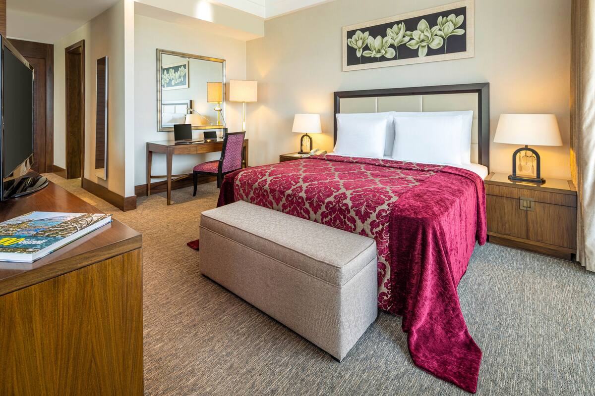 A hotel room with a large bed, a desk with a chair, a television, and a bedside table with a lamp.