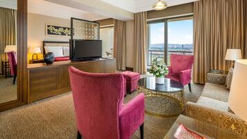 A hotel room with a large window, a flat-screen TV, a sofa, two armchairs, a coffee table, and a decorative vase.