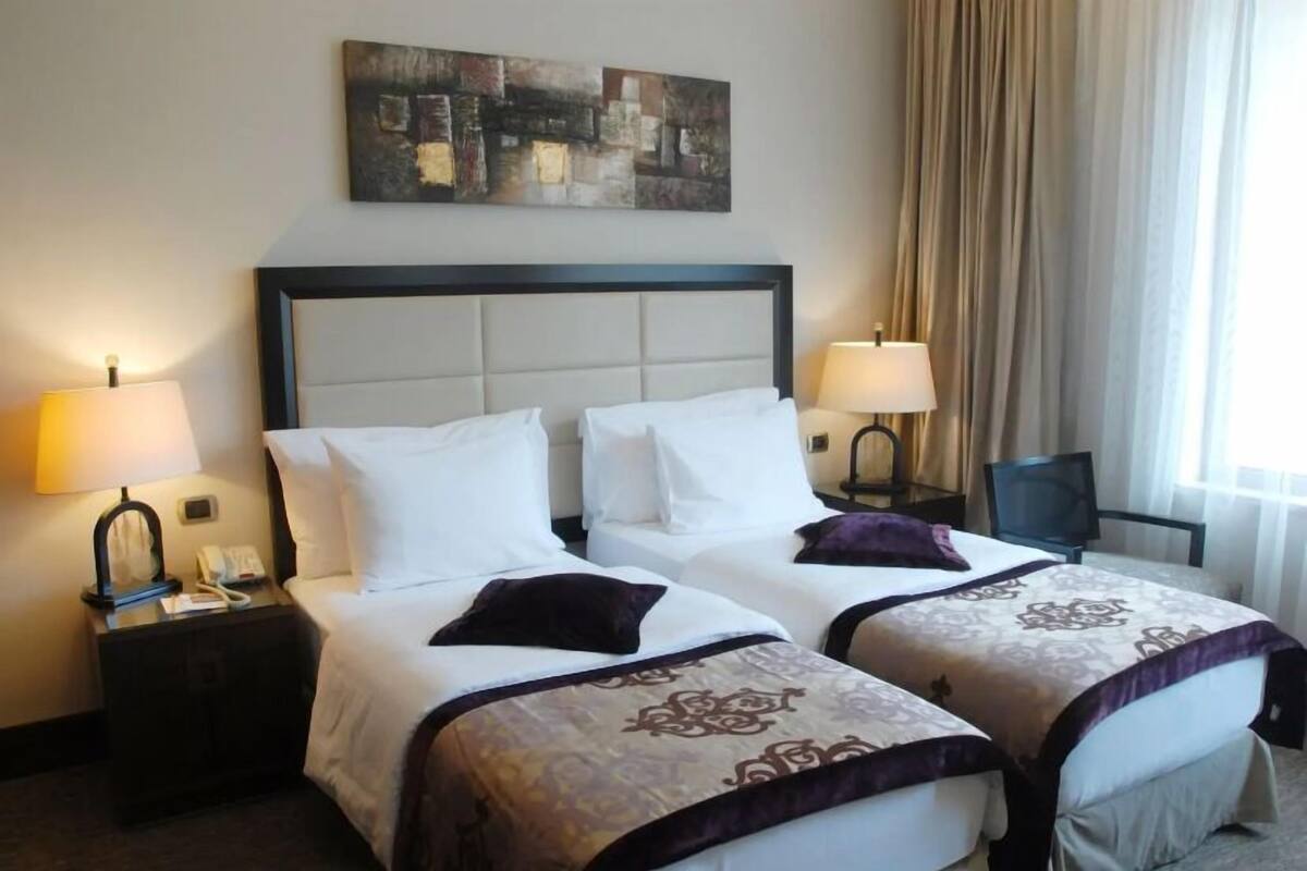 A hotel room with two beds, a headboard, bedside lamps, a chair, a painting on the wall, and a window with curtains.