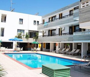 A pool area with lounge chairs and umbrellas, a building with balconies, and a clear blue sky. - Agrino Hotel Apartments (Ayia Napa)
