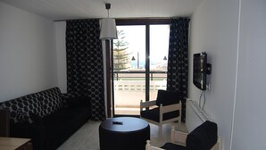 A living room with a sofa, a round ottoman, a chair, and a television mounted on the wall. - Agrino Hotel Apartments (Ayia Napa)