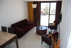 Een woonkamer met een bank, een rond salontafeltje, een eettafel en stoelen. Er is een groot raam met gordijnen.
