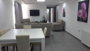 A modern living room with a sofa, a dining table with chairs, a TV mounted on the wall, and a potted plant. - Agrino Hotel Apartments (Ayia Napa)