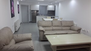 A modern living room with a beige sofa, a matching armchair, and a wooden coffee table. A kitchen with a refrigerator and a dining table is visible in the background. - Agrino Hotel Apartments (Ayia Napa)