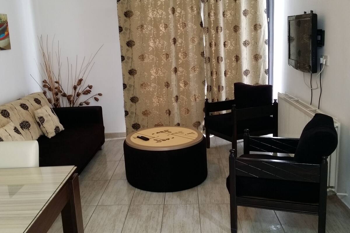 A living room with a sofa, a round coffee table, a dining table, and a TV mounted on the wall.