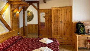 Un dormitorio con techo de madera, una cama con colcha roja estampada, un armario de madera y un escritorio con una mochila encima.